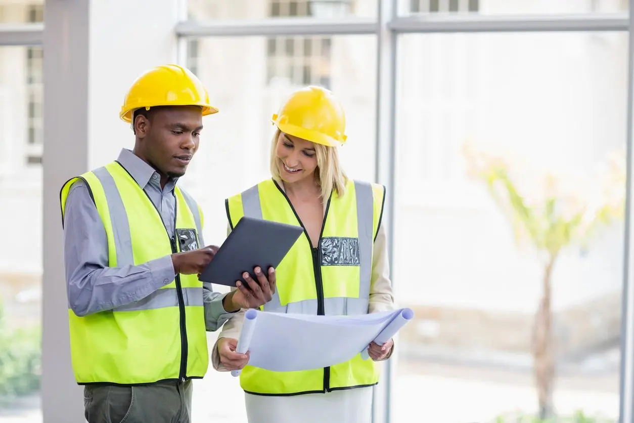 Two people in yellow vests and hard hats looking at a tablet.