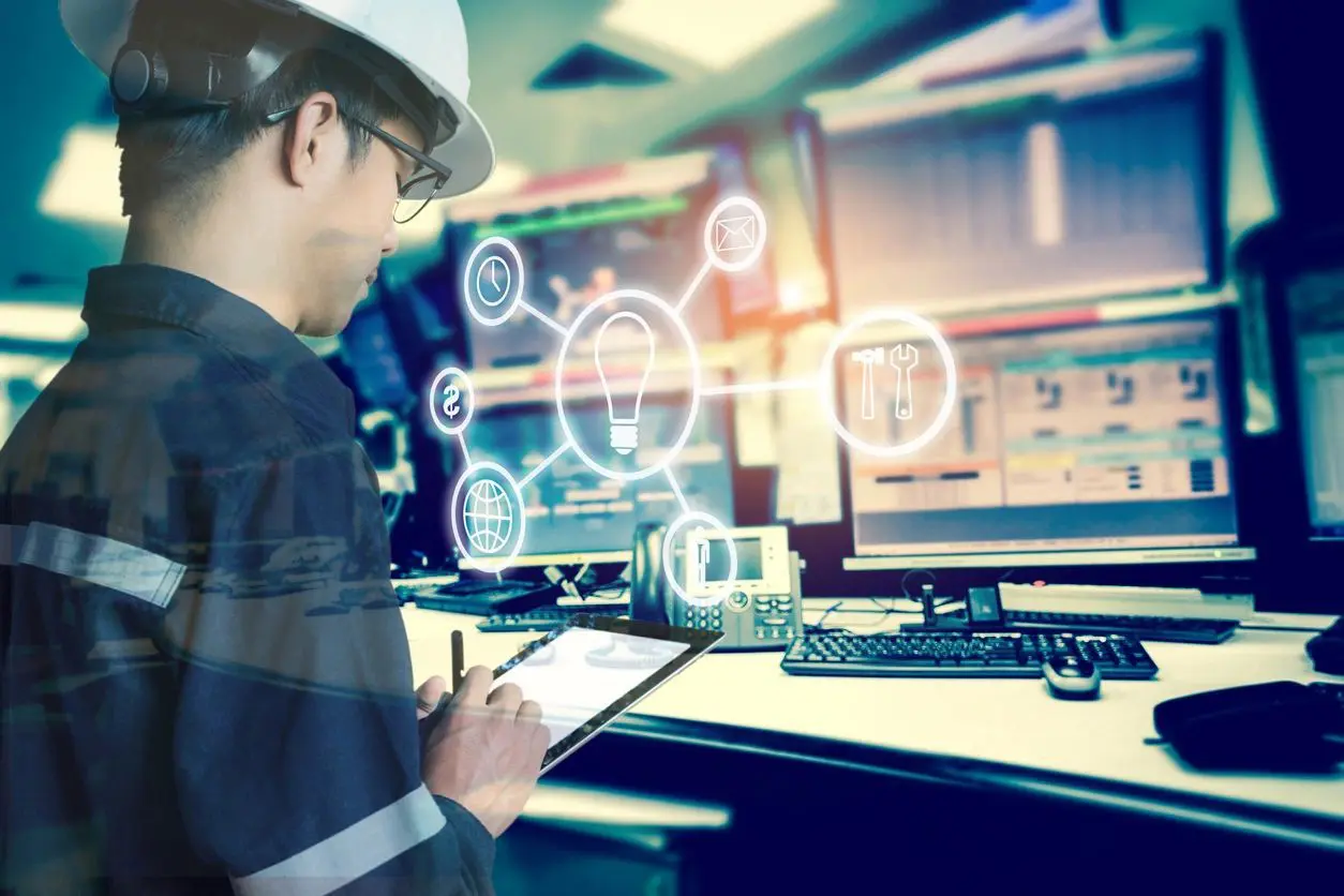 A man in hard hat writing on paper near computer monitors.